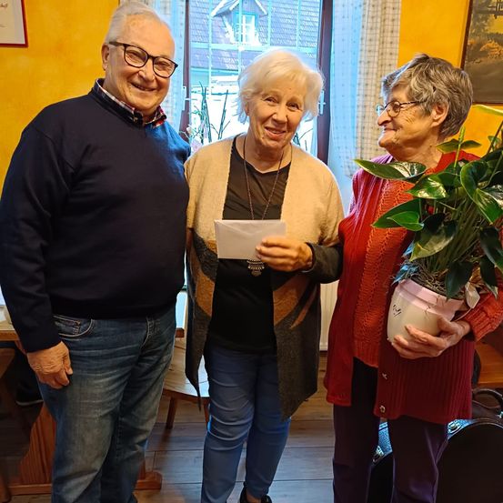 Drei ältere Personen sind drinnen, lächeln und posieren für ein Foto. Die Frau in der Mitte hält ein Stück Papier und eine Halskette. Die Frau auf der rechten Seite hält eine Topfpflanze. Hinter ihnen ist ein Fenster mit Vorhängen.