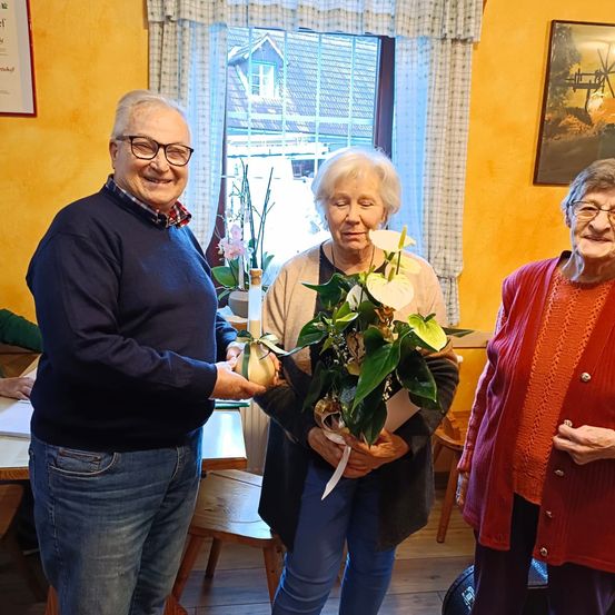 Drei ältere Personen sind drinnen, ein Mann mit Brille überreicht einer Frau in der Mitte einen Blumenstrauß. Die Frau hält die Blumen, während eine weitere Frau rechts von ihr steht. Ein Fenster und ein Bilderrahmen befinden sich hinter ihnen.