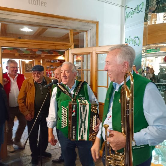 Zwei Männer in grünen Westen, einer spielt Akkordeon und der andere hält eine Trompete, stehen neben einem Mikrofon in einem Restaurant. Hinter ihnen steht ein Mann in einer braunen Jacke und Hut mit einem anderen Mann, der eine Brille trägt.