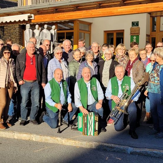 Eine Gruppe von Menschen steht vor einem Gebäude und posiert für ein Foto. Einige tragen Brillen und halten Musikinstrumente. Statuen sind hinter ihnen, und ein grünes Schild ist an der Wand.