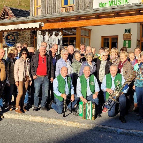 Eine Gruppe von Menschen posiert für ein Foto vor einem Gebäude, einige mit Musikinstrumenten. Das Gebäude hat ein Schild mit der Aufschrift 'der MUSIK'.