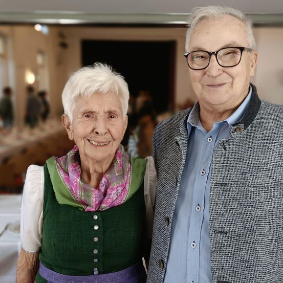 Ein älteres Paar steht eng beieinander in einem Raum und lächelt. Die Frau trägt ein grünes Kleid mit einem Schal. Der Mann trägt eine Brille und eine Jacke. Hinter ihnen sind verschwommene Figuren und Tische zu sehen.