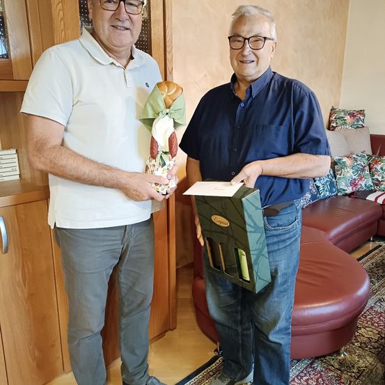 Zwei Männer stehen in einem Wohnzimmer, einer hält ein verpacktes Geschenk und eine Weinschachtel, während der andere ein Stück Papier in der Hand hat.