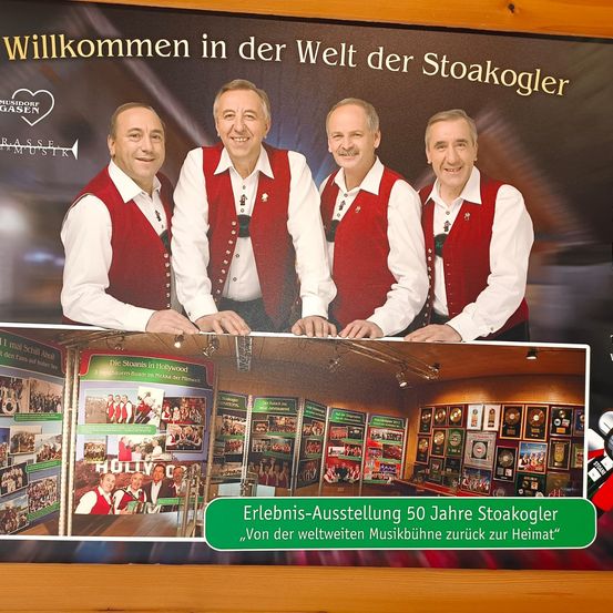 Plakat von vier Männern in roten Westen mit dem Text 'Willkommen in der Welt der Stoakogler'. Fotos und Auszeichnungen darunter mit einem grünen Banner 'Erlebnis-Ausstellung 50 Jahre Stoakogler'.