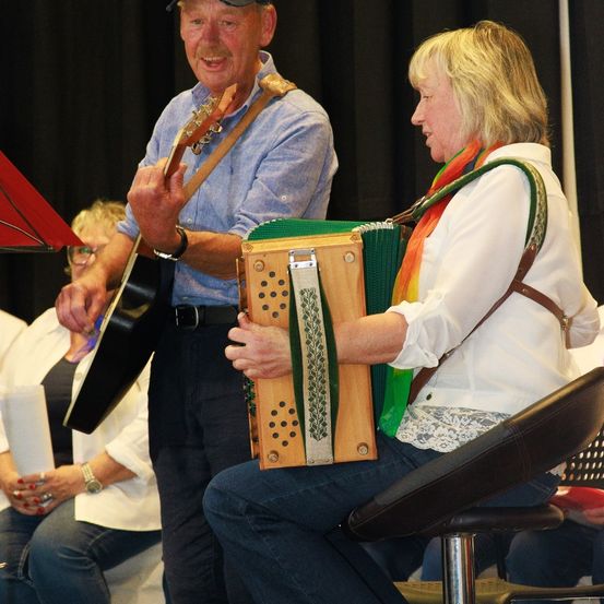 Ein älterer Mann und eine ältere Frau spielen Instrumente, vielleicht bei einem Konzert. Der Mann spielt Gitarre und die Frau Akkordeon. Sie lächeln beide. Dahinter sitzen andere Menschen und schauen zu.
