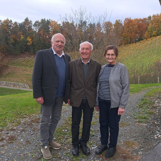 Drei Personen, zwei Männer und eine Frau, stehen auf einem Kiesweg und lächeln für ein Foto, während ein Weinberg im Hintergrund zu sehen ist.