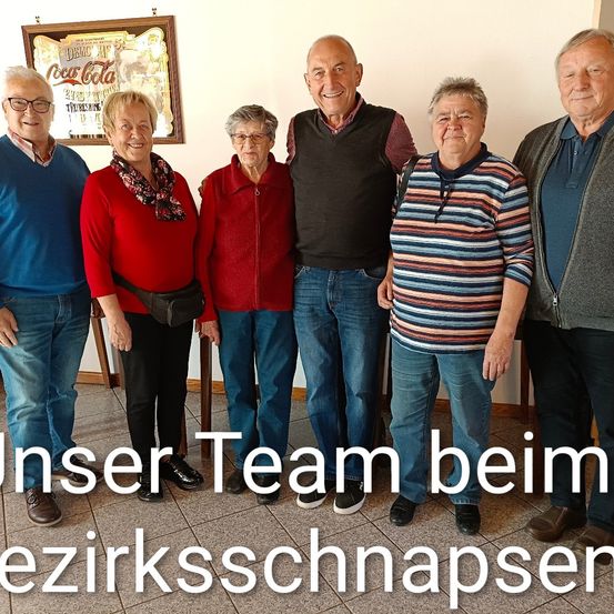 Sechs Menschen stehen eng beieinander mit Lächeln auf den Gesichtern. Der Text 'Unser Team beim ezirksschnapsen' befindet sich am unteren Rand des Bildes.