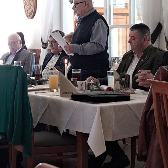 Eine Gruppe älterer Männer sitzt um einen Tisch in einem Restaurant. Ein Mann steht und hält ein Papier, und eine Kerze ist auf dem Tisch angezündet.