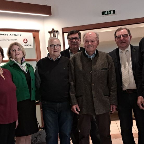 Eine Gruppe von sechs Personen, darunter drei Männer und drei Frauen, steht zusammen in einem Raum. Sie alle lächeln und posieren für ein Foto. Der Mann ganz rechts trägt eine Krawatte und einen Gürtel. Der Mann auf der linken Seite trägt eine Jacke. Der Mann in der Mitte trägt eine Brille.