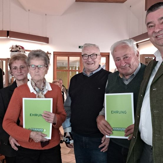 Fünf Personen stehen eng beieinander und lächeln. Sie halten grüne Broschüren mit dem Titel 'Ehrun'. Die Umgebung ist innen, mit weißen Wänden, Holztüren und Lampen, die von der Decke hängen.