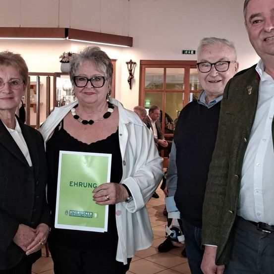 Vier Menschen, drei Frauen und ein Mann, lächeln für die Kamera. Die Frau in der Mitte hält ein grünes Schild mit weißem Text. Dahinter befindet sich ein Raum mit Holzwänden und einer Lampe.