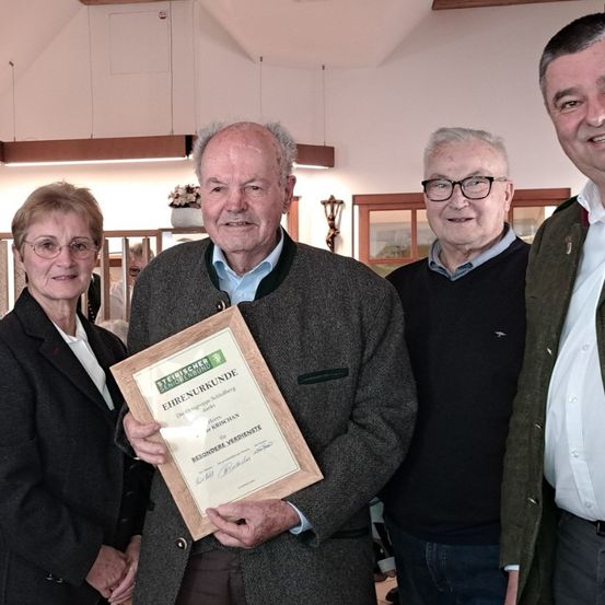 Ein älterer Mann hält eine gerahmte Urkunde, umgeben von drei anderen Personen. Zwei Männer und eine Frau sind formell gekleidet. Der Mann rechts trägt eine grüne Jacke. Hinter ihnen ist eine weiße Wand mit Bilderrahmen.