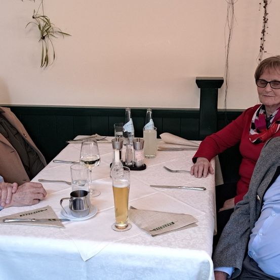 Drei Personen sitzen lächelnd an einem Tisch. Eine Frau in Rot trägt eine Brille, ein Mann in einem braunen Mantel sitzt links von ihr und ein anderer Mann in einem blauen Hemd rechts von ihr. Gläser und eine Tasse stehen auf dem Tisch.