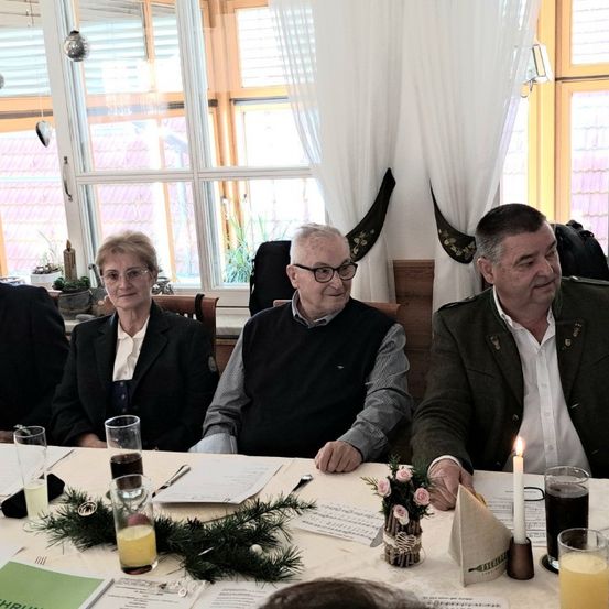 Eine Gruppe von Menschen sitzt um einen Tisch herum, lächelt und schaut in die Kamera. Sie sind von verschiedenen Gegenständen umgeben, darunter eine Kerze, Blumen und Gläser mit Saft. Der Mann rechts trägt eine Militäruniform.