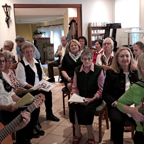 Eine Gruppe älterer Frauen sitzt gemeinsam in einem Raum, einige lesen aus Büchern, andere spielen Gitarre. Hinter ihnen sitzt ein Mann mit einem Glas Saft. Ein Kruzifix und eine Topfpflanze sind im Hintergrund zu sehen.