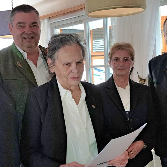 Eine Gruppe von Menschen steht in einem Raum mit einem Fenster im Hintergrund. Eine ältere Frau liest ein Papier, flankiert von zwei anderen, während ein Mann neben ihr lächelt.