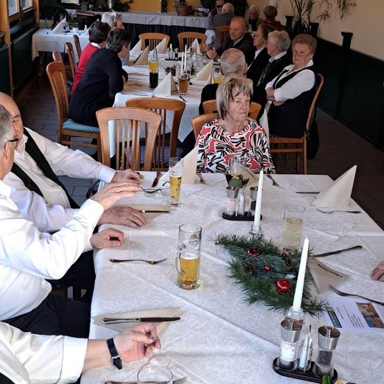 Mehrere Personen sitzen um einen langen Tisch in einem Restaurant. Sie führen ein Gespräch. Der Tisch ist mit Getränken, Servietten und Besteck gedeckt. Es gibt Weihnachtsdekorationen auf dem Tisch.
