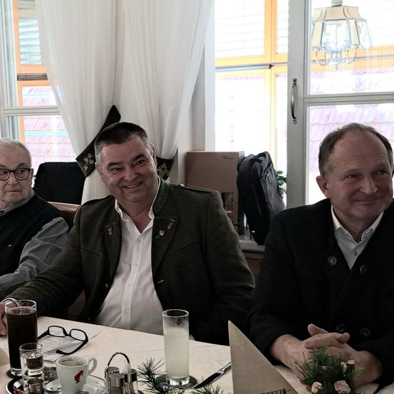 Drei Männer sitzen an einem Tisch und lächeln für ein Foto. Der Mann in der Mitte trägt eine grüne Jacke, die anderen schwarz. Hinter ihnen ist ein Fenster mit einem Kronleuchter zu sehen, und Vorhänge sind gezogen.