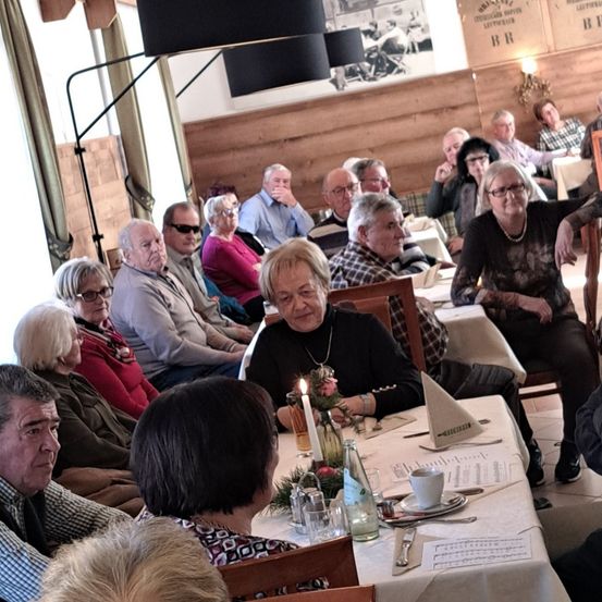 Eine Gruppe älterer Menschen versammelt sich in einem Restaurant. Eine Frau sitzt an einem Tisch mit einer brennenden Kerze, einem Blumenvase und einer Speisekarte.
