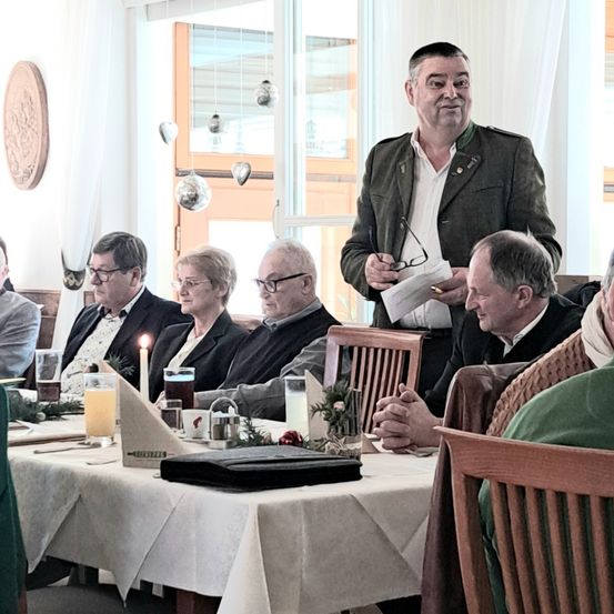 Mehrere Personen sitzen um einen Tisch herum in einem Innenraum. Ein Mann steht hinter ihnen und hält ein Papier und eine Brille. Gläser, Kerzen und Tassen stehen auf dem Tisch. Ein Fenster mit Vorhängen und Dekorationen befindet sich hinter ihnen.