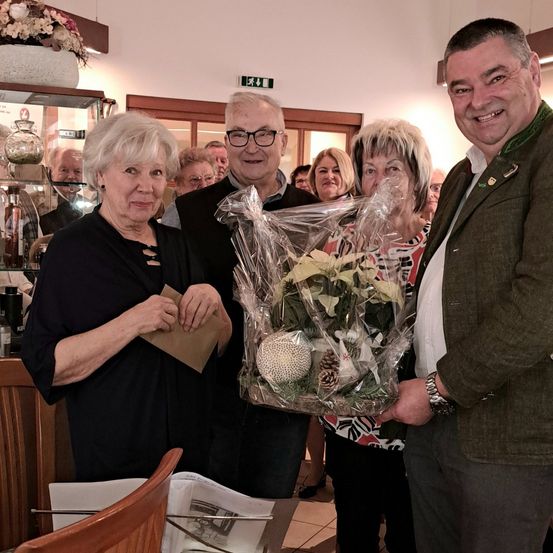 Mehrere Menschen stehen in einem Raum, zwei von ihnen halten ein Blumenarrangement. Eine Frau in einem schwarzen Oberteil hält ein Stück Papier. Ein Mann in einem grünen Jackett lächelt. Im Hintergrund ist ein Regal mit verschiedenen Gegenständen zu sehen.