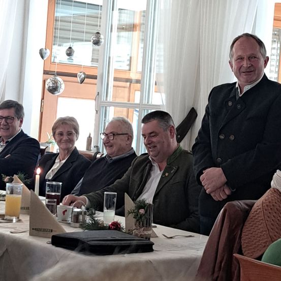 Eine Gruppe von Menschen sitzt an einem Tisch. Ein Mann mit Brille steht hinter ihnen. Auf dem Tisch stehen eine Kerze, ein Glas Saft und eine kleine Pflanze. Es gibt ein Fenster mit weißen Vorhängen und einigen dekorativen Gegenständen, die daran hängen.