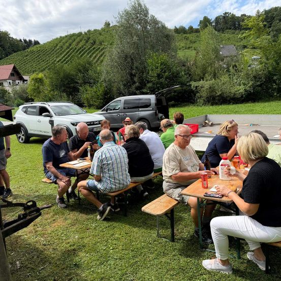 Eine Gruppe von Menschen sitzt an Picknicktischen in einem Grasbereich, mit geparkten Autos dahinter und einem Weinberg im Hintergrund.