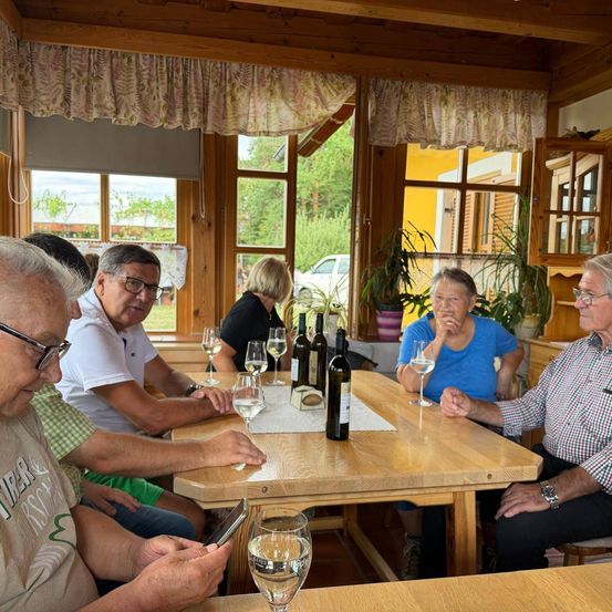 Eine Gruppe von Menschen sitzt um einen Holztisch in einem rustikalen Raum und genießt Wein und Gespräche. Flaschen und Gläser stehen auf dem Tisch, und Pflanzen und ein Auto sind durch die Fenster zu sehen.