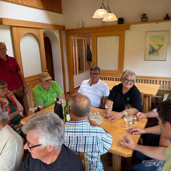 Eine Gruppe von Menschen sitzt um einen Tisch herum, einige halten Weingläser in der Hand, wahrscheinlich in einem Essbereich. Flaschen und Gläser stehen auf dem Tisch.