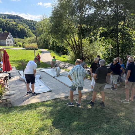Eine Gruppe von Menschen versammelt sich an einem sonnigen Tag um einen Minigolfplatz, wobei eine Person spielt und andere zuschauen.