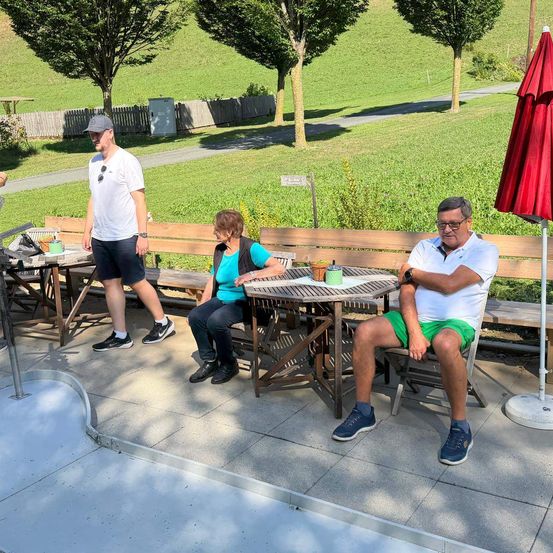 Drei Menschen sitzen auf einer Parkbank. Ein Mann steht vor ihnen. Vor ihnen steht ein Tisch mit einer Tasse darauf. Rechts steht ein roter Schirm.