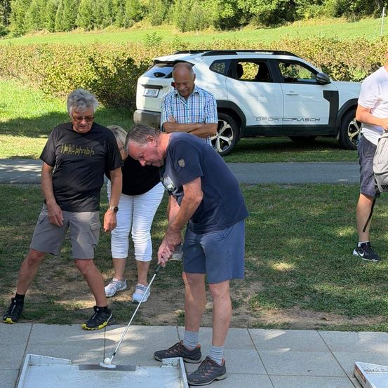 Eine Gruppe von Menschen spielt Golf auf einem Betonweg. Ein Mann beugt sich vor, um den Ball zu schlagen. Hinter ihm beobachtet ein Mann ihn. Eine Frau steht hinter ihm. Ein Auto ist hinter ihnen geparkt.