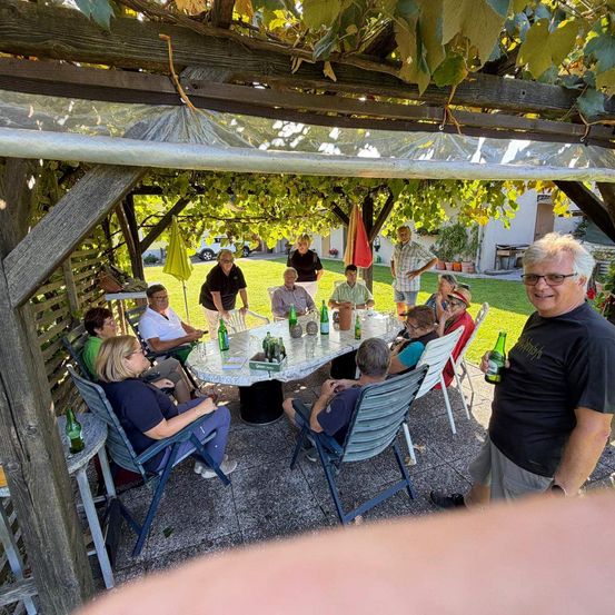 Eine Gruppe von Menschen sitzt um einen Tisch in einem Garten, einige halten Flaschen. Ein Mann steht vor ihnen und lächelt.