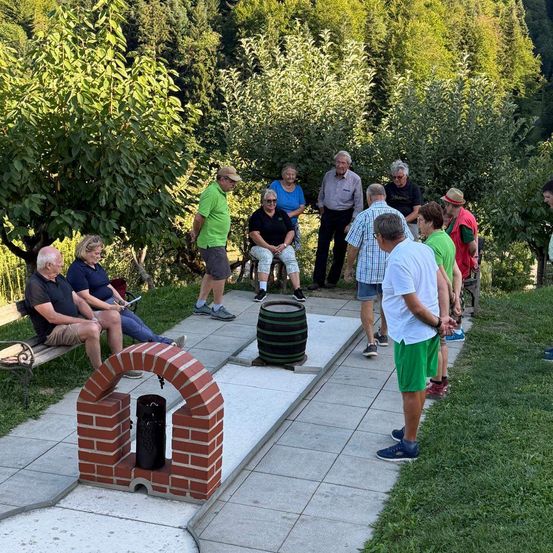 Eine Gruppe von Menschen versammelt sich um einen Minigolfplatz in einem Garten. Einige stehen, während andere auf einer Bank sitzen. Ein Fass ist als Ziel auf dem Boden platziert.