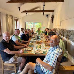 Eine Gruppe von Menschen sitzt an einem langen Tisch und genießt eine Mahlzeit. Der Tisch ist mit Tellern, Gläsern und verschiedenen Speisen gedeckt. Der Raum hat Holzbalken und Steinwände.