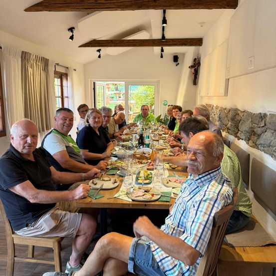 Eine Gruppe von Menschen sitzt an einem langen Tisch und genießt eine Mahlzeit. Der Tisch ist mit Tellern, Gläsern und verschiedenen Speisen gedeckt. Der Raum hat Holzbalken und Steinwände.