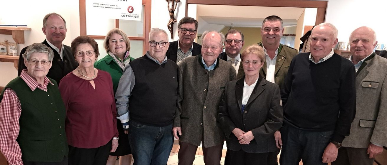 Eine Gruppe von Menschen, darunter Männer und Frauen, posiert für ein Foto. Sie stehen alle lächelnd eng beieinander. Ein Kreuz hängt an der Wand hinter ihnen.