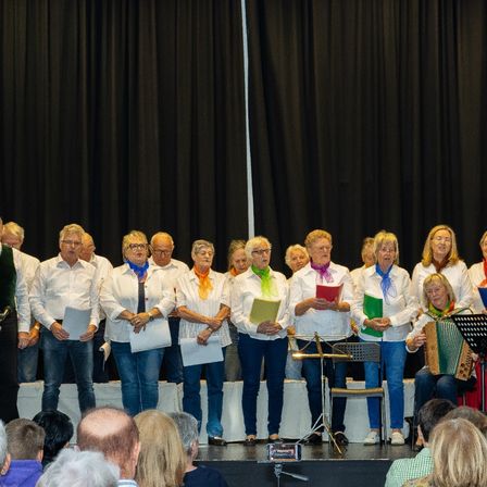 Ein Chor tritt auf der Bühne auf und spielt verschiedene Instrumente, darunter ein Akkordeon. Die Künstler tragen weiße Hemden und Schals. Hinter ihnen befindet sich ein schwarzer Vorhang.