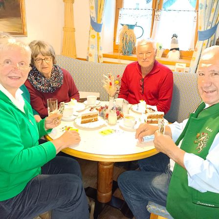 Vier Personen sitzen um einen Tisch mit Kuchen, Getränken und Servietten. Der Mann auf der rechten Seite trägt eine grüne Schürze. Der Raum hat ein Fenster mit Vorhängen und einem Bilderrahmen.
