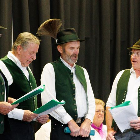 Drei Männer in grünen Westen und Hüten stehen auf der Bühne, halten Papiere und singen, mit einer Frau in Brille und Schal im Hintergrund.