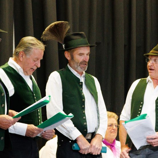 Drei Männer in grünen Westen und Hüten stehen auf der Bühne, halten Papiere und singen, mit einer Frau in Brille und Schal im Hintergrund.