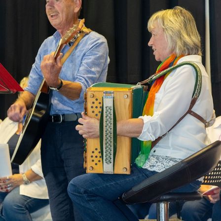 Ein älterer Mann spielt Gitarre, während eine Frau neben ihm Akkordeon spielt. Sie treten auf der Bühne auf, und hinter ihnen sitzen einige Leute und schauen zu.