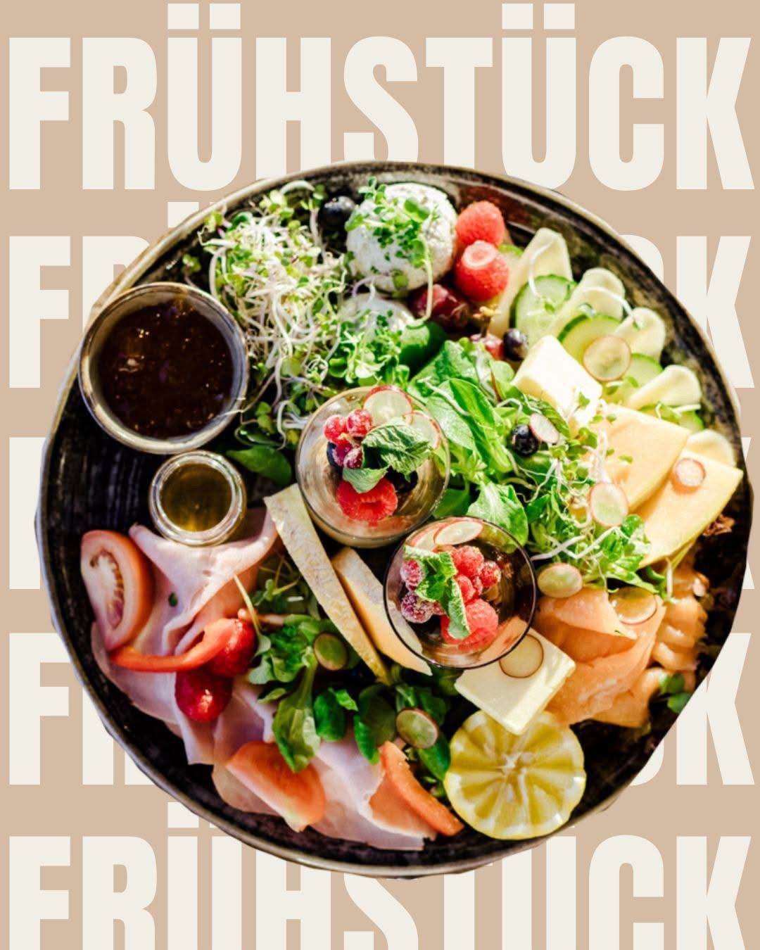 Ein runder Teller gefüllt mit einer Vielfalt frischer Lebensmittel, darunter Scheiben von Käse, Tomaten, Gurken und eine Schüssel mit Beeren. Es sind auch Gewürze wie Olivenöl und Honig vorhanden. Der Hintergrund ist beige mit weißem Text, der 'FRUITSTOCK' lautet.