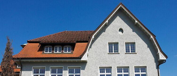 Ein modernes Wohngebäude mit einem roten Dach und mehreren Fenstern. Das Gebäude ist aus grauem Stein und vor ihm steht ein Baum. Der Himmel ist klar und blau.