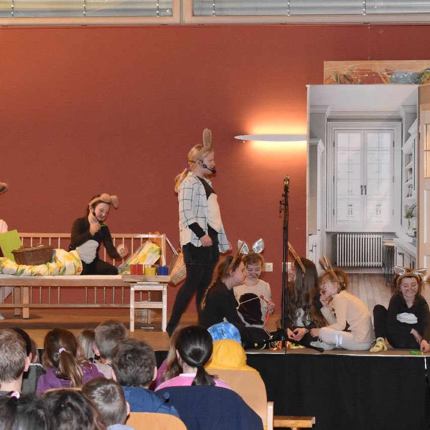 Eine Gruppe von Kindern führt ein Theaterstück auf einer Bühne auf. Die Bühne ist mit einem Bett, Stühlen und einer Tür dekoriert. Die Kinder tragen Hasenohren und Gesichtsbemalung.