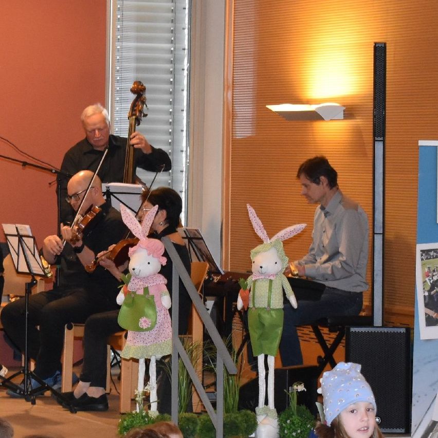 Eine Gruppe von Musikern spielt Instrumente, darunter Cello, Geige und Klavier, in einem Raum mit Bühne. Sie werden von zwei Kaninchen-Maskottchen begleitet, von denen eines eine Karotte hält.