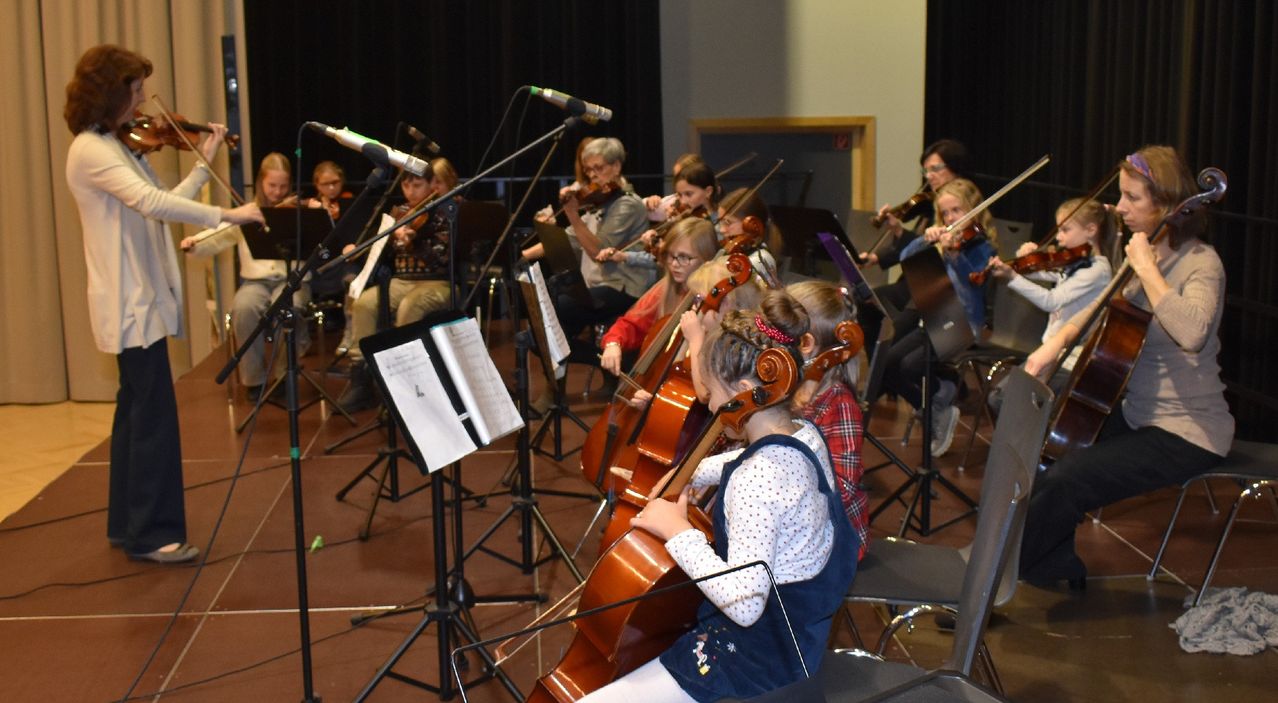 Eine Gruppe junger Musiker, darunter Geiger und Cellisten, übt in einem Raum mit Notenständern und Mikrofonen. Sie tragen alle Brillen und einige haben Turnschuhe an.