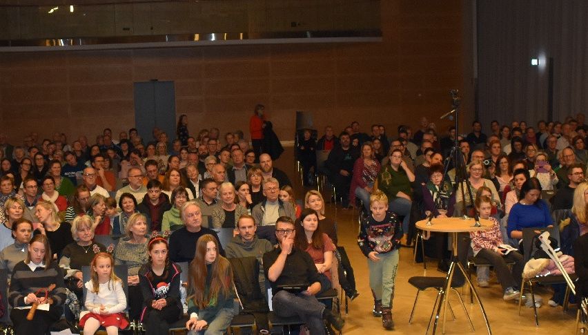 Ein großes Publikum versammelt sich in einem Theater, gefüllt mit verschiedenen Personen, die sitzen und stehen, mit einer Bühne und einem Mikrofon vorne aufgebaut.