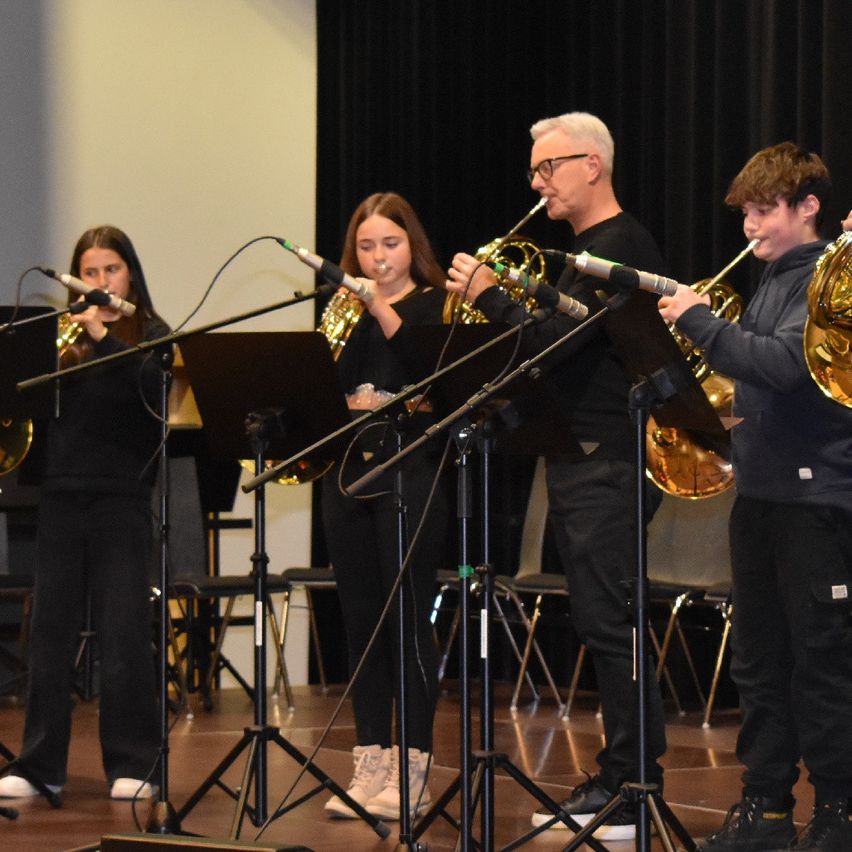 Fünf Musiker, darunter zwei Mädchen und ein älterer Mann, spielen in einem Konzertsaal französische Hörner mit Mikrofonen an Ständern.