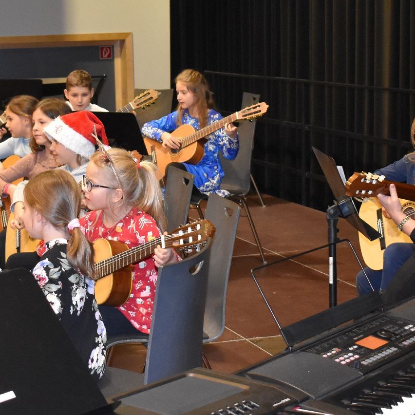 Mehrere Kinder sitzen auf Stühlen und spielen Gitarren in einem Musikraum, eines trägt einen Weihnachtsmannhut. Ein Klavier ist im Vordergrund sichtbar.